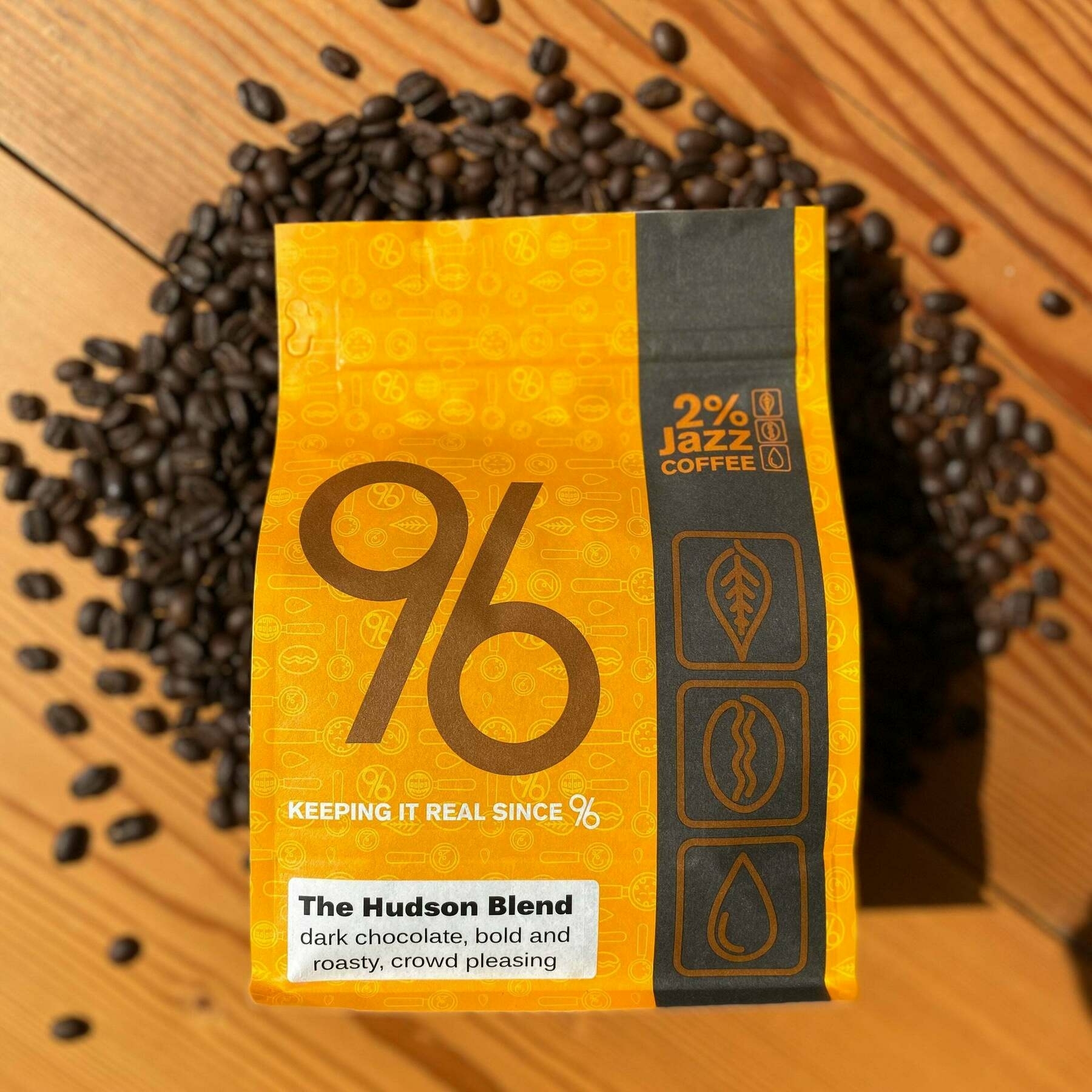 A bag of 2% Jazz Coffee's "The Hudson Blend" is surrounded by scattered coffee beans on a wooden surface.
