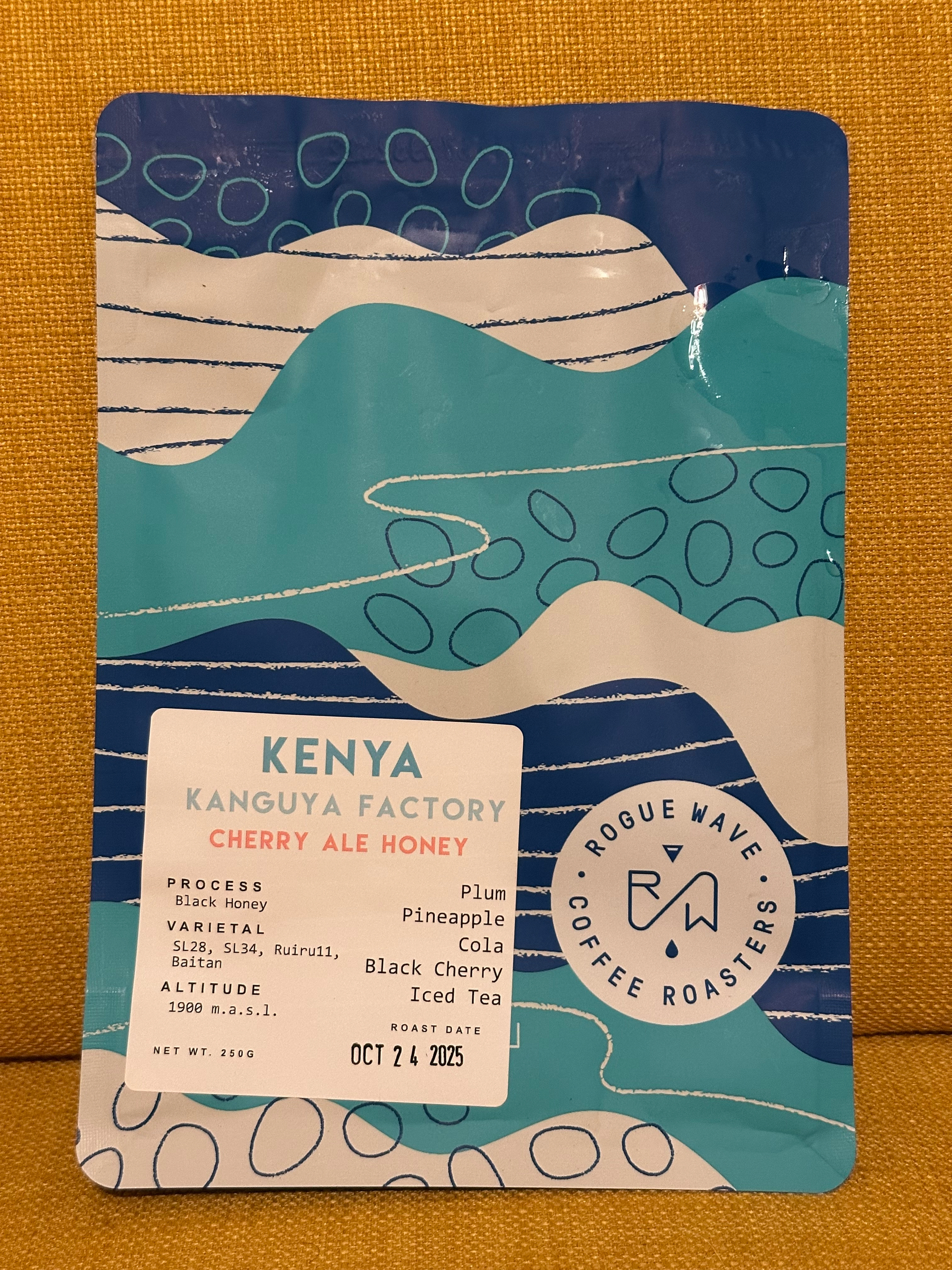 A coffee bag labeled Kenya Kanguya Factory Cherry Ale Honey with tasting notes of plum, pineapple, cola, black cherry, and iced tea, designed in shades of blue and white.
