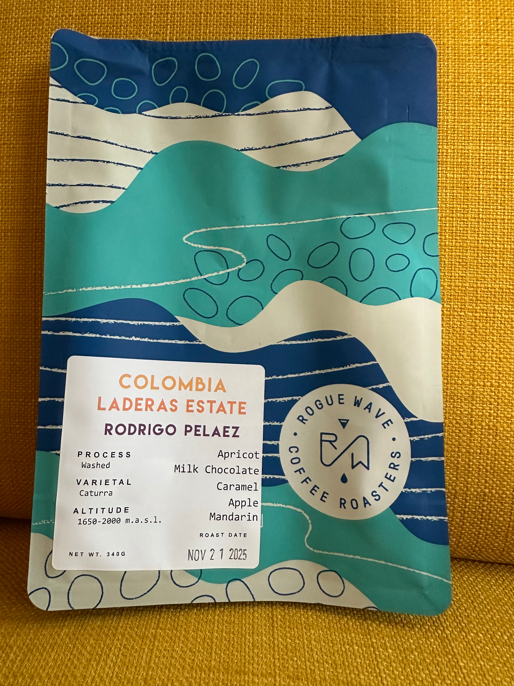 A colorful coffee bag with decorative patterns features details about Colombia Laderas Estate coffee, including flavors like apricot and milk chocolate, and a roast date of November 21, 2025.