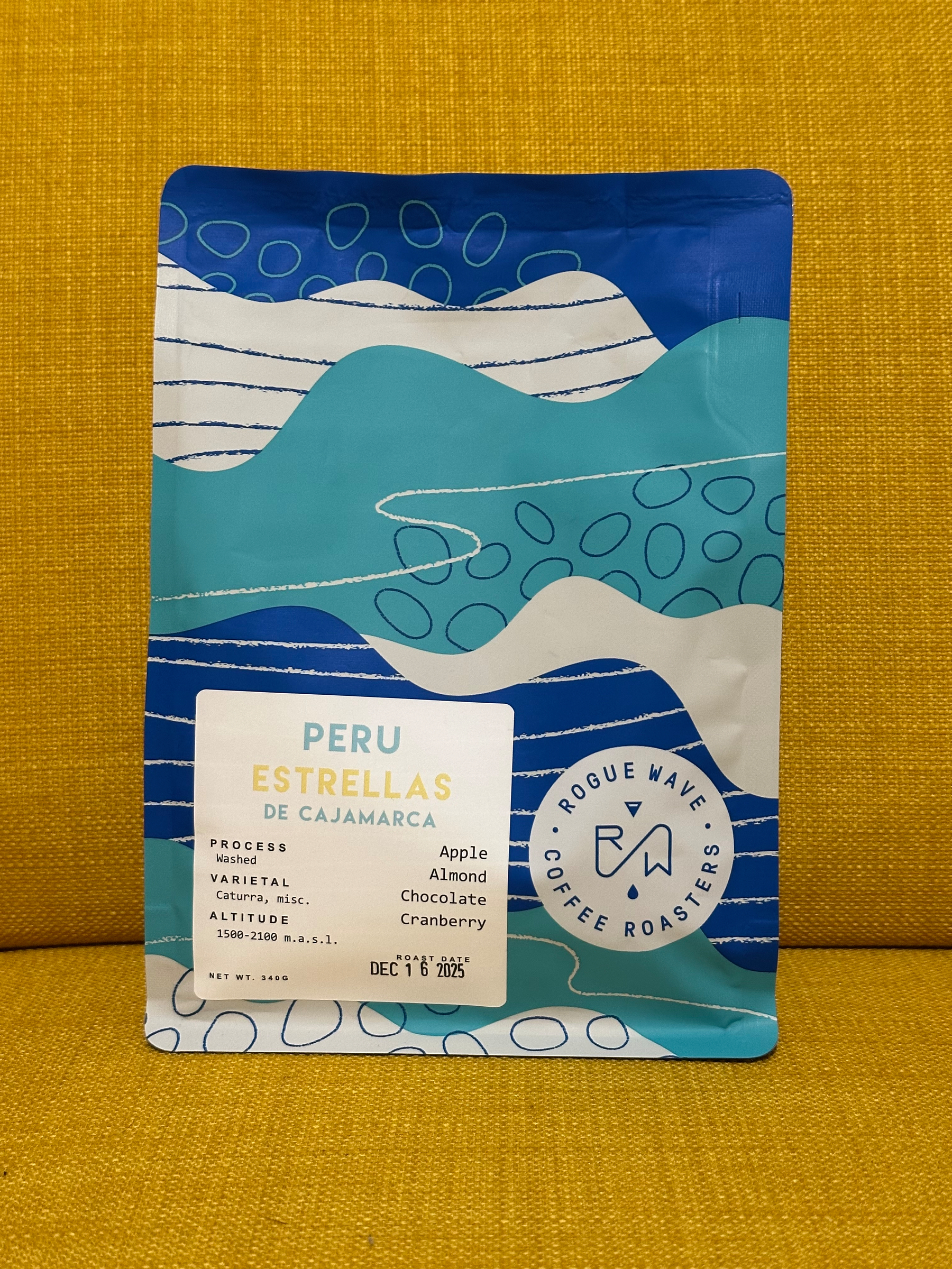 A colorful coffee packaging featuring information about the Peru Estrellas de Cajamarca coffee, including tasting notes of apple, almond, chocolate, and cranberry, with a bold design and the Rogue Wave Coffee Roasters logo.