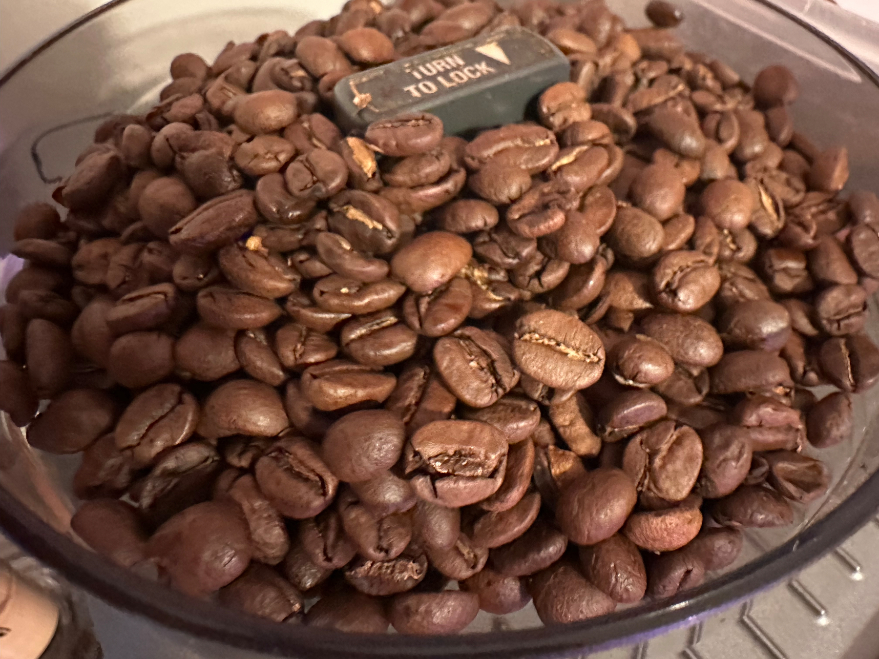 A container filled with roasted coffee beans has a small label or dial displaying the text Turn to Lock.