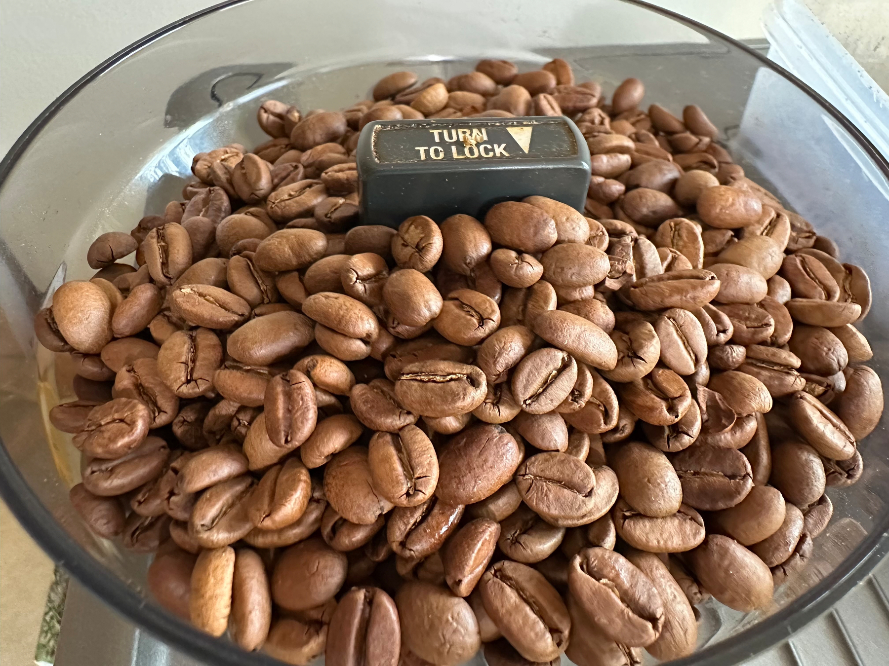 A container filled with roasted coffee beans has a black locking mechanism on top labeled TURN TO LOCK.