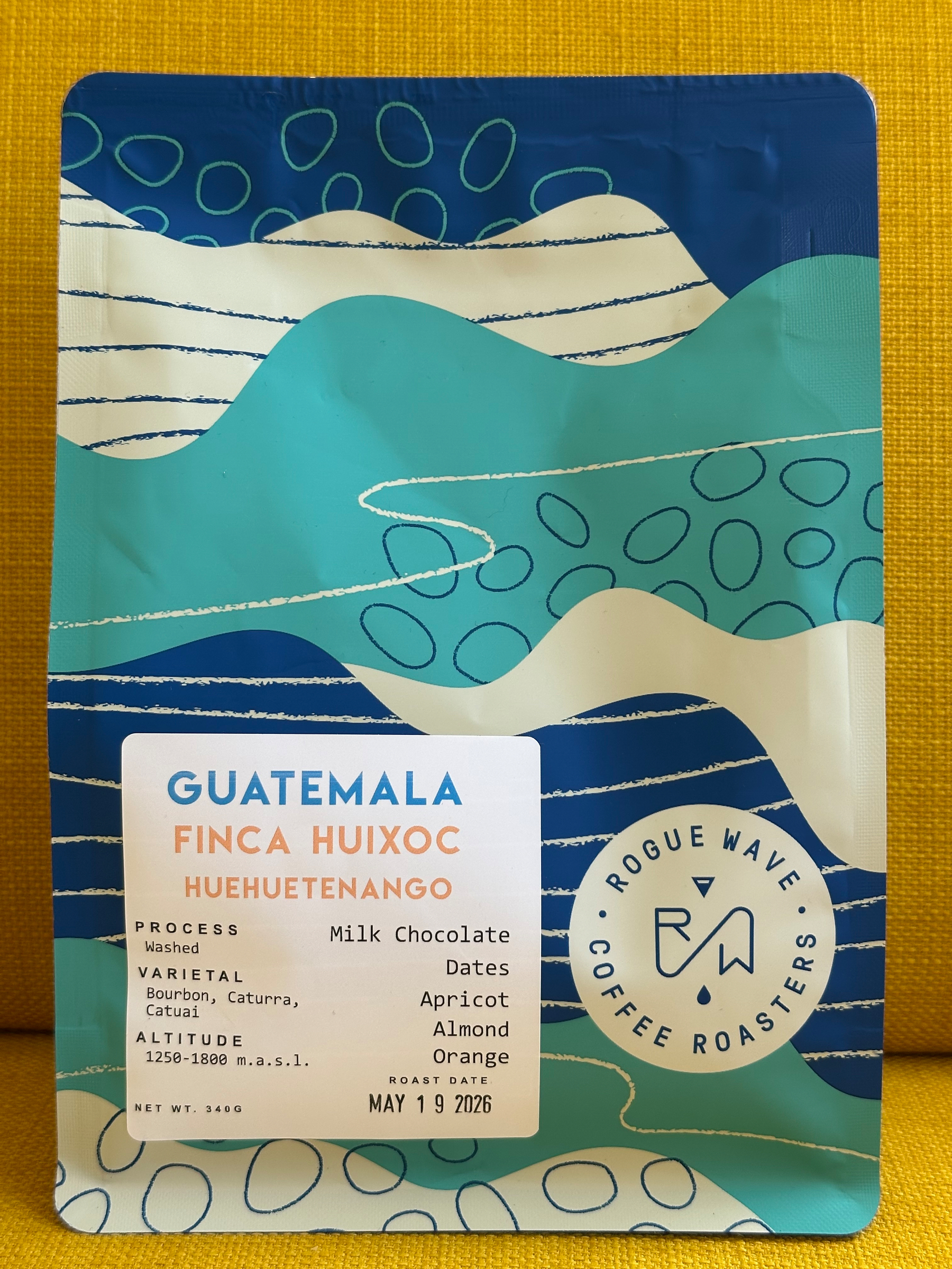 A colorful coffee bag features abstract blue and green designs with details about the coffee origin from Guatemala and flavor notes like milk chocolate, dates, apricot, almond and orange.