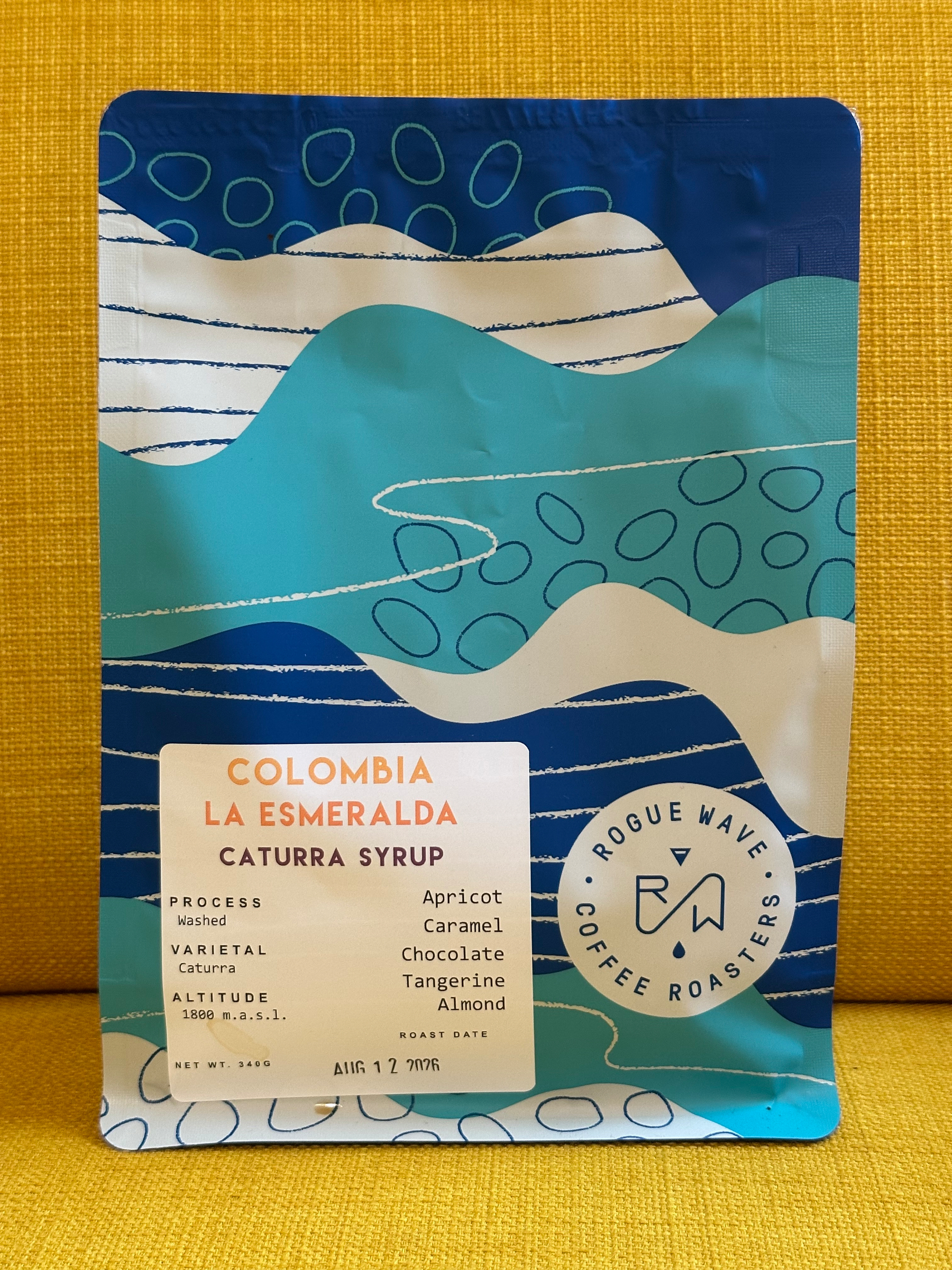 A colorful blue and white bag of Colombia La Esmeralda Caturra Syrup coffee from Rogue Wave Coffee Roasters is displayed on a yellow background.