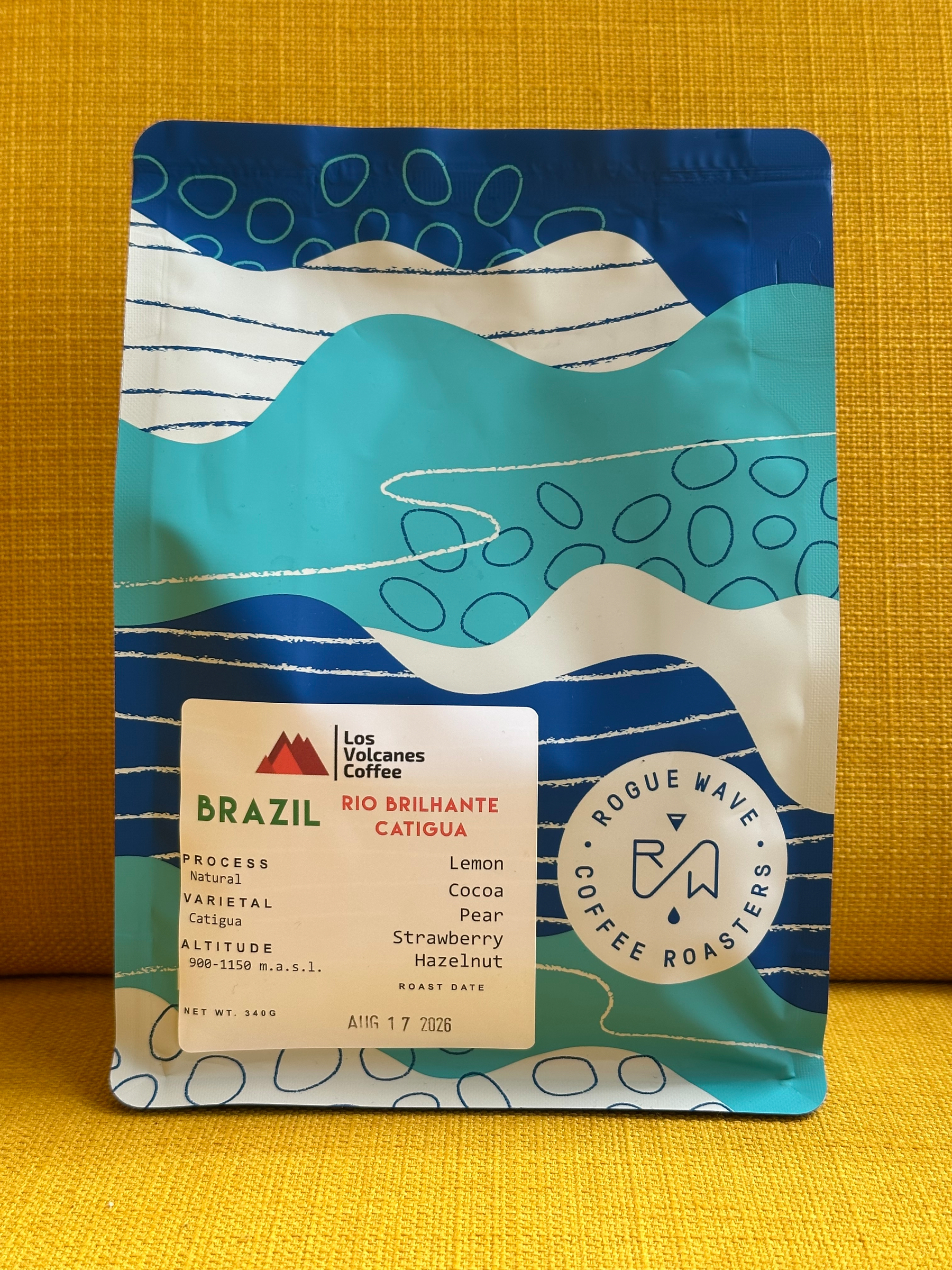 A colourful coffee bag labeled Brazil Rio Brilhante Catigua from Rogue Wave Coffee Roasters, featuring flavour notes of lemon, cocoa, pear, strawberry, and hazelnut, is placed against a yellow background.