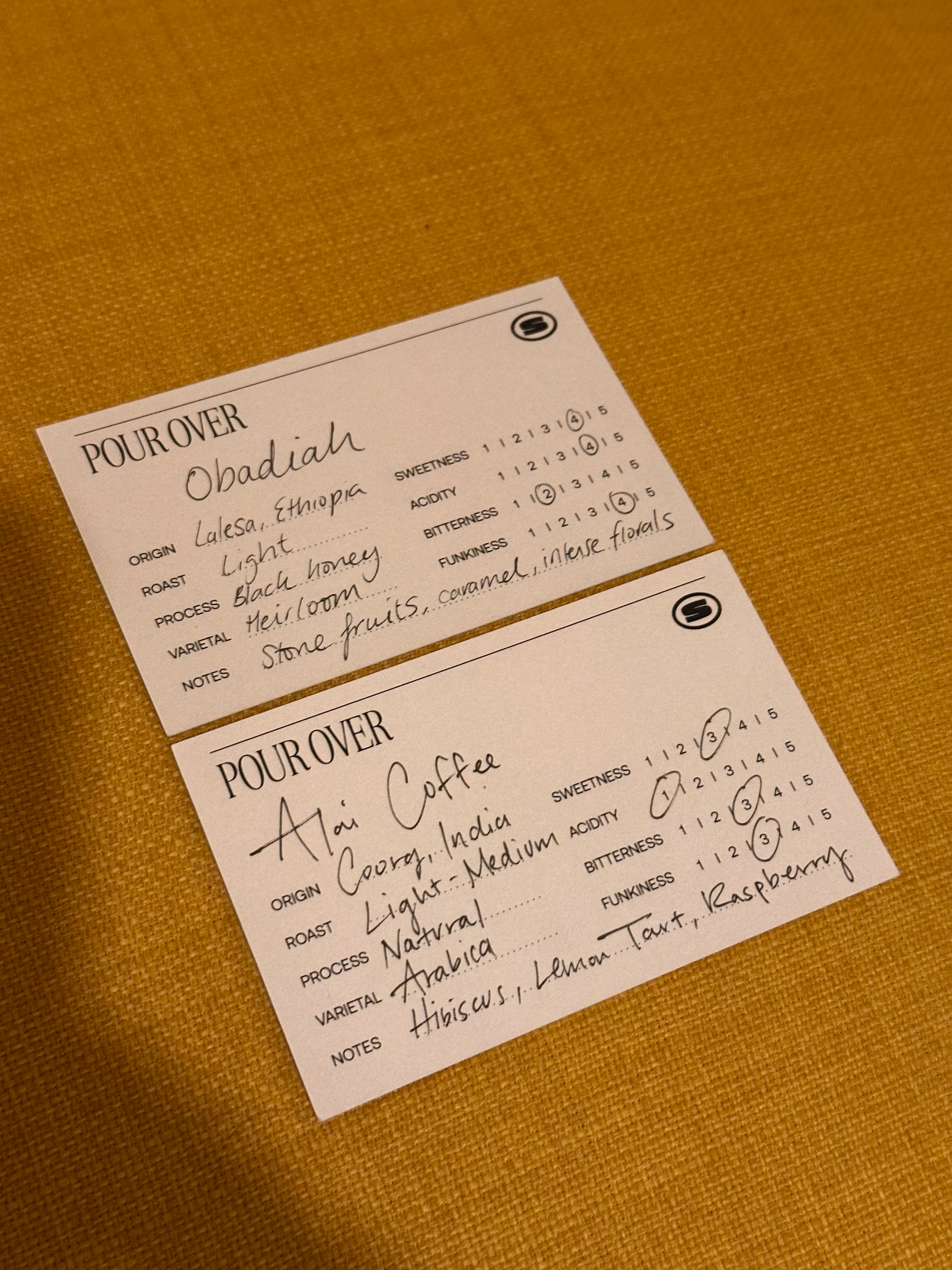 Two pour-over coffee tasting notes cards show details about the origin, roast, process, varietal, taste notes, and ratings for sweetness, acidity, bitterness, and funkiness for Obadiah from Ethiopia and Aoi Coffee from India.